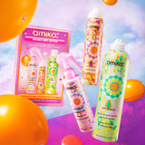 A promotional image for Amika's 'Ready, Set, Style' set, featuring the products displayed against a vibrant background with floating orange balloons. The products, including Brooklyn Bombshell Blowout Spray, Perk Up Dry Shampoo, and Undone Volume and Matte Texture Spray, are shown outside the box, floating in mid-air. The packaging box is also visible on the left, showcasing the products inside. The overall design is colorful and energetic, with a playful, retro-inspired aesthetic.  